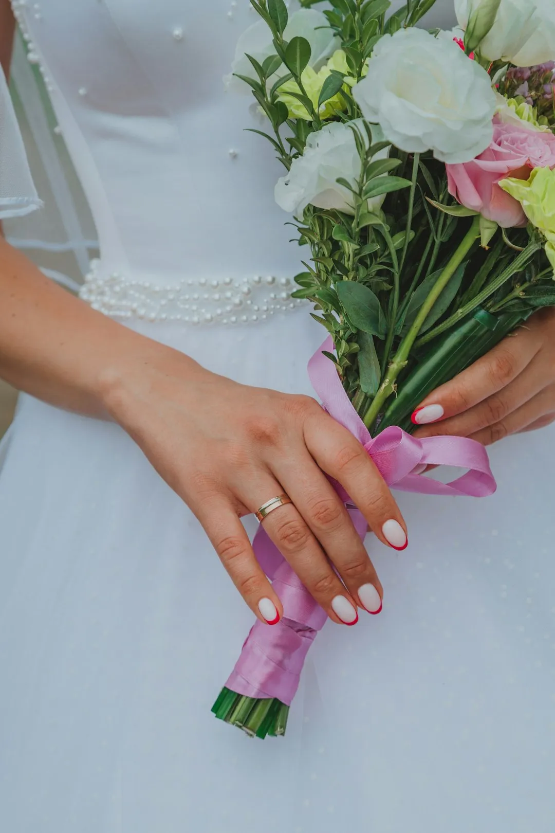 A bride holds a bouquet of flowers with pink ribbon and displays her french manicure with red tips