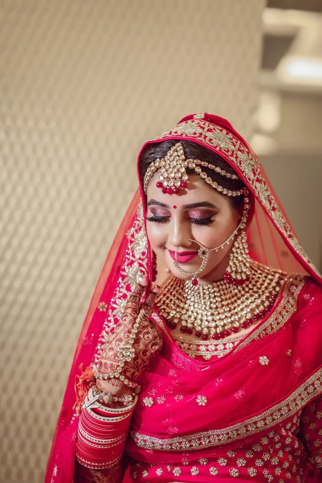 A bride wearing heavy gold jewelry, a red headscarf, and red bangles looks down with her eyes closed