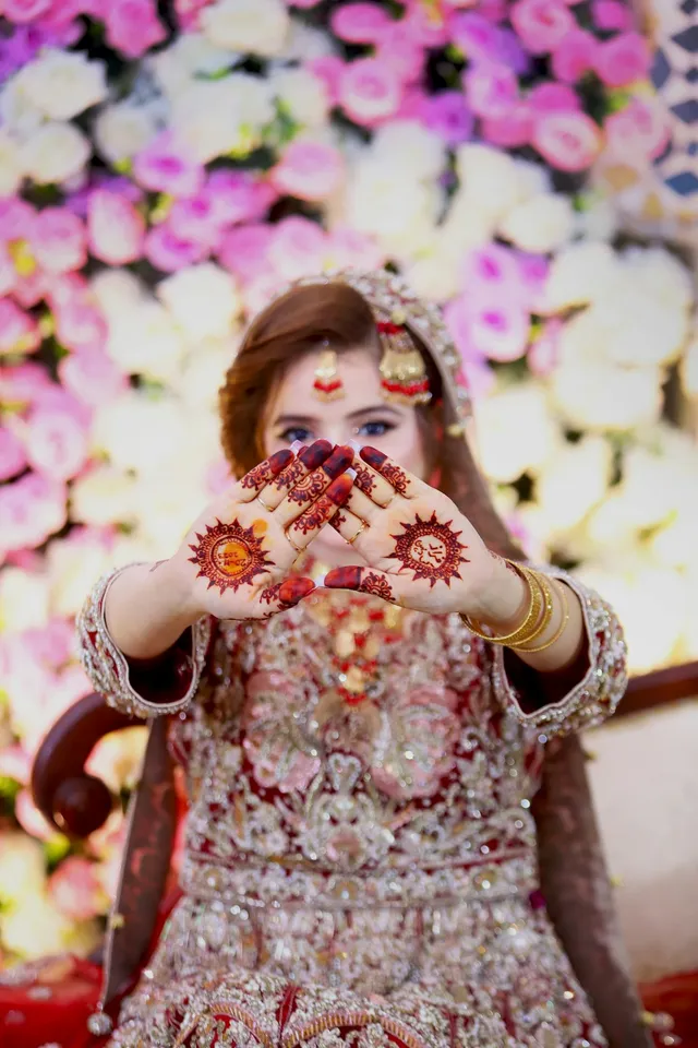 Mehndi bride showing henna-covered palms in front of a pink and white floral backdrop