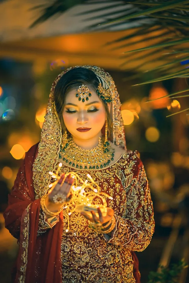 A woman in ornate bridal attire and jewelry looks down at glowing string lights held gently in her hands